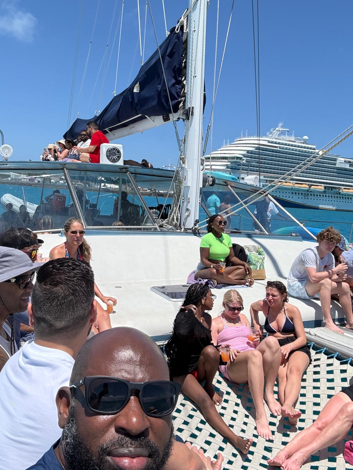 Guests relaxing on the deck of a catamaran beside a cruise ship on a sunny day